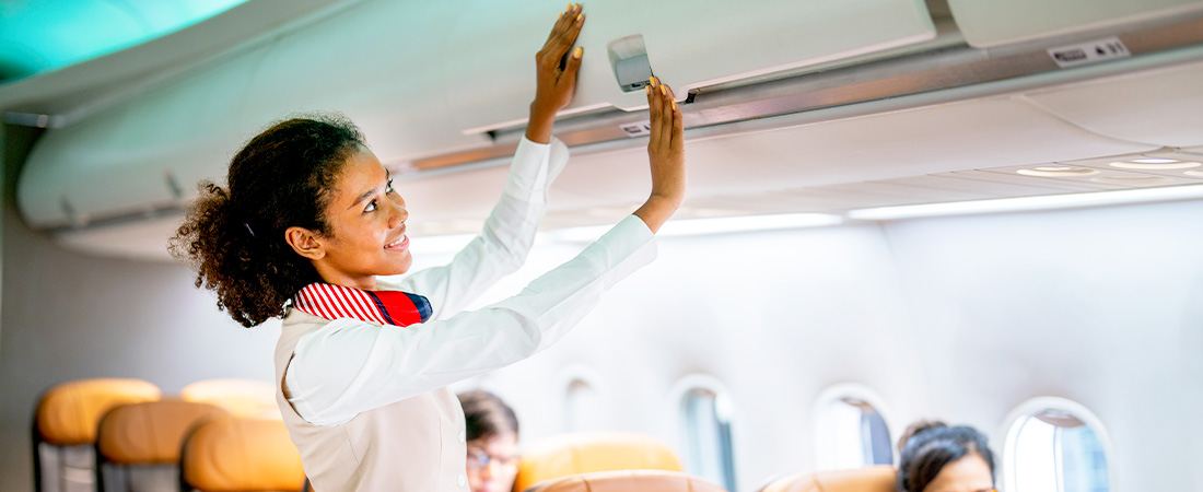 Una azafata (TCP) con uniforme blanco y pañuelo rojo cierra un compartimento de equipaje superior dentro de la cabina de un avión.